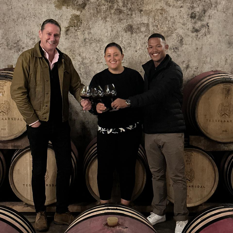 Three people stand with wine glasses in front of barrels stamped with the word Tesselaarsdal