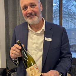 A middle-aged man in a jacket is holding a bottle of wine with a sepia-tinted label